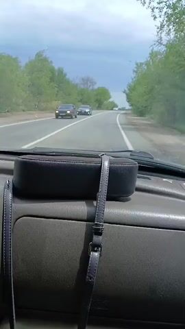 The video shows a first-person view from inside a car driving on a paved road. The road is lined with trees and greenery on both sides. The sky is cloudy. In the distance, there are several other cars on the road, some in the same lane and some in the oncoming lane. A dark-colored purse with straps is visible on the dashboard in the foreground.