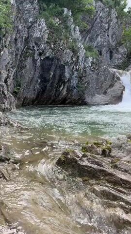 The video shows a small waterfall cascading into a pool of water, surrounded by rocky terrain and greenery. The water is clear and flowing rapidly, with small ripples and currents visible. The rocks are mostly gray and covered with patches of moss and vegetation. The scene is set in a natural environment, likely a forest or mountainous area. The sky is overcast, providing soft, diffused lighting.