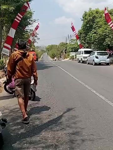 The video shows a street scene with several people and vehicles. A man in a brown jacket and shorts stands next to a motorcycle on the side of the road, facing away from the camera. Another person is walking on the road further down, in the distance. Several flags, red and white, are hanging across the road. Cars and vans are parked or driving on the road. The environment appears to be a rural or semi-urban area with trees and buildings visible on either side of the road. The weather is sunny.