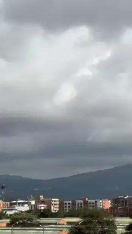 The video shows a cloudy sky with white and dark grey clouds. At the bottom of the frame, there is a row of buildings and some trees. In the background, mountains are visible, partially obscured by the clouds. The clouds change from mostly dark grey to patches of blue sky with fluffy white clouds.