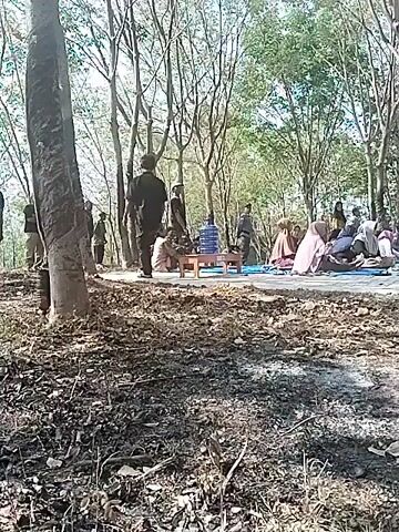 A group of people are gathered outdoors in a wooded area. Many are seated on the ground, some on blue mats. Some individuals are wearing headscarves. In the background, there is a building with a pitched roof and a veranda. Several large black speakers are stacked on top of each other. Trees with bare branches are visible throughout the scene. Some areas on the ground appear to be scorched or burnt.