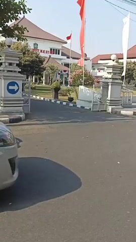The video opens with a clear view of a building with a sign reading "Kantor Wilayah". There are flags, one red and one white, flying in front of the building. A black Mercedes-Benz sedan is parked on the side of the road, partially obscuring the view of the building. The road appears to be paved and is lined with a curb. Orange traffic barriers are visible on the right side of the frame. The sky is clear and blue, suggesting a sunny day.