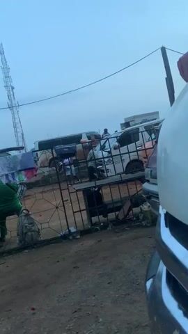 The video shows a busy outdoor area, likely a marketplace or transport hub, with several vehicles including vans and a bus. A tall communications tower is visible in the background. A metal fence separates the foreground from the area with the vehicles and people. Several people are gathered, some sitting on benches or sitting by the fence. Clothing is hanging on a clothesline near the fence. A large backpack is on the ground by the fence. The ground appears to be dirt or gravel, and there is some debris scattered around. The sky is overcast and the light suggests it is either early morning or late afternoon. A white vehicle is partially visible in the foreground, taking up a significant portion of the right side of the frame. The overall impression is one of a lively, somewhat disorganized, public space.