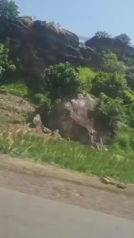 The video shows a natural landscape with rocky hills covered in green vegetation and trees. The camera appears to be moving, possibly from inside a vehicle, as indicated by the blurred foreground and the perspective of the passing scenery. The sky is clear and blue. There are no people or animals visible. The terrain is uneven and appears to be a natural, undeveloped area.
