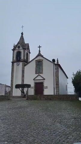 The video shows a church on an overcast day. The church is white with gray stone trim. It has a steeple with a cross on top and a clock on the side. In front of the church is a small, well-trimmed tree. The ground is paved with gray stones. A green bush is to the right of the church. In the background, other buildings and a fence can be seen.