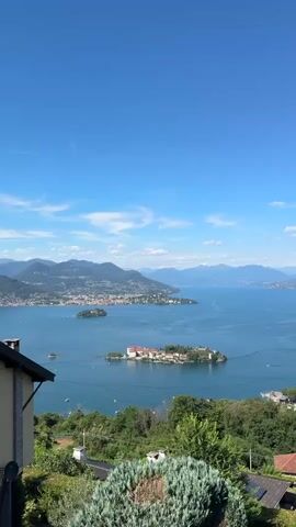 This is a vertical video with a clear blue sky and scattered clouds. The video captures a panoramic view of a large body of water, likely a lake, with islands and a distant mountainous landscape. In the foreground, lush green trees and bushes dominate, with glimpses of rooftops of houses nestled among the foliage. As the video progresses, the camera pans to the right, revealing a town or city built along the shoreline, with buildings clustered together and extending up the hillsides. The water is calm, with small boats visible on its surface. The overall impression is one of a scenic and serene natural environment, possibly a vacation destination, with the architecture of the town harmonizing with the surrounding landscape. There are no people visible in the video.