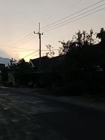 A person wearing a black helmet and jacket is riding a motorcycle on a paved road at dusk. They are silhouetted against a pale orange sky with visible power lines and utility poles. Several houses and trees are visible on the side of the road. Another motorcycle passes in the opposite direction. The license plate on the first motorcycle reads AE 6769 P1.