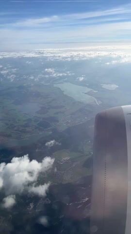 An aerial video captures a panoramic view from an airplane window over Vils, Austria, during the daytime. The right side of the frame features a portion of the airplane's engine, reflecting sunlight. Below, a vast landscape unfolds with vibrant green fields, several distinct lakes, and winding waterways, indicating an agricultural and hydrographical region. Mountains rise in the distance, partially covered by clouds, and small towns with clusters of buildings are visible along the lake shores and within the valleys. The sky above is a clear blue with scattered white clouds, while a layer of fluffy clouds floats below the plane's altitude, creating a soft, ethereal backdrop. The overall scene suggests bright, sunny weather with excellent visibility. No people or text are visible.