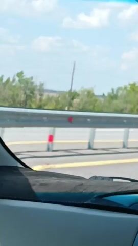 The video shows a view from inside a car driving on a highway. The camera is pointed forward, capturing the road ahead and the surrounding landscape. A metal guardrail runs parallel to the highway, with reflective markers attached to its posts. Beyond the guardrail, there are trees and vegetation, and in the distance, a utility pole and a white sign with red markings are visible. The sky is blue with some scattered clouds. The dashboard of the car is partially visible in the foreground.
