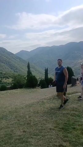 A group of people are walking on a grassy hill. In the foreground, a man in a blue tank top and black shorts walks to the right, holding a phone. Behind him, a woman in a pink shirt and denim shorts walks to the left, also holding a phone. In the background, there are rolling green mountains under a partly cloudy sky. Several trees are scattered across the landscape, and some ancient stone structures are visible. The people appear to be tourists exploring the area.