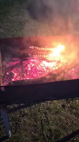 The video shows a close-up of a fire burning in a metal fire pit. The fire pit is filled with glowing embers and burning wood, emitting smoke. The embers glow a bright red color. The fire pit is sitting on a grassy area.  A faint song is audible in the background.
