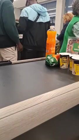 A static shot depicts two individuals in casual attire at a supermarket checkout lane, with a variety of groceries placed on the conveyor belt. The person on the left is wearing a dark green hooded sweatshirt and light-colored shorts. They are examining something in their hand, possibly a phone or wallet. The other person is dressed in a black jacket with a light blue hood, black pants, and a cap. A black cross-body bag is visible on their back. 

The grocery items on the conveyor belt include a large bottle of orange juice, a bag of chips, and several containers labeled "Sabor Pollo Frango". The conveyor belt itself is a flat, gray surface with a light-colored wooden edge.

Other individuals can be seen further back in the line, including a person wearing a green vest, likely a supermarket employee. The background is illuminated by overhead lighting, typical of a retail environment, and includes glimpses of supermarket aisles and shelving.