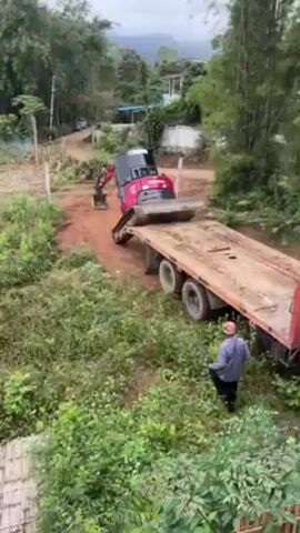A red and gray Yanmar mini excavator is carefully driving up onto a flatbed trailer in a rural area. A man wearing a plaid shirt and a safety helmet stands near the trailer, apparently directing the excavator operator. The trailer is attached to a red truck, and the ground around the truck is muddy and unpaved. Lush green vegetation surrounds the area, with trees and shrubs visible in the background. A house and other buildings are situated further up the hill. The sky is overcast.