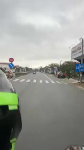 A person rides a motorcycle on a highway on an overcast day. The road has two lanes in each direction, separated by a concrete barrier. Houses and trees are visible on either side of the road. Other vehicles are on the road.