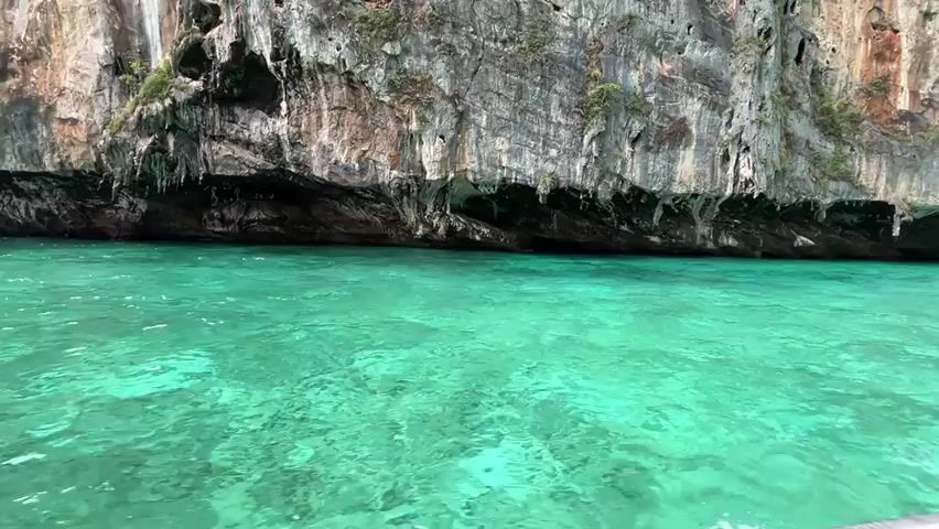 A person is on a boat moving through turquoise water near a sheer, rocky cliff face. The water is clear and reflects the light, showing the ripples and texture of the sea. The cliff is tall and imposing, with dark crevices and overhangs. Small patches of green vegetation grow on the upper parts of the cliff. The sky is visible in the distance, and other boats can be seen further along the coast. The perspective is low, close to the water's surface.