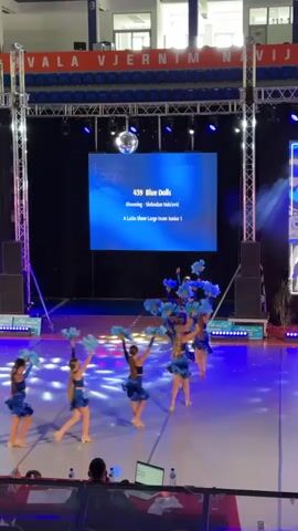 The video shows a dance performance on a stage. There are several young girls in blue costumes, holding blue props, dancing in sync. A large screen behind them displays text that reads "459 Blue Dolls" and other performance details. The stage is lit with blue and purple lights, and there is smoke effect. In the background, there are spectators sitting in bleachers. The overall environment appears to be an indoor arena or gymnasium.