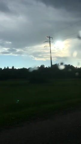 This video depicts a view from a moving vehicle, likely a car, traveling along a road. The foreground shows a dark road surface and a metallic guardrail, suggesting the vehicle is on a paved road, possibly a highway or a country road.

The background reveals a rural landscape. A dark line of trees forms a dense forest or treeline across the mid-ground. Beyond the trees, there are patches of green, suggesting fields or meadows. The sky is dramatic, with a mix of dark, heavy clouds and brighter, sunlit areas. The sun is visible, peeking through the clouds, creating a diffused and somewhat hazy light. Several bright, circular orbs are scattered across the frame, which appear to be water droplets or smudges on the vehicle's window, indicating it might be raining or has recently rained.

The time of day appears to be late afternoon or early evening, given the quality of light and the mix of sun and clouds. The weather is changeable, with indications of recent or ongoing precipitation. The scene evokes a sense of travel and contemplation, with the changing sky and the passing landscape. There are no visible people or text in the media. The setting is a natural, outdoor environment in Russia, specifically Tutaev.