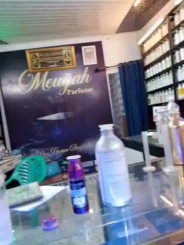 The video shows two men in a shop that sells perfumes. The shop is well-lit and has many shelves filled with bottles of perfume. The man on the left is wearing a black t-shirt and jeans. He is holding a phone in his left hand and appears to be paying the man on the right. The man on the right is wearing a black t-shirt with a pattern and is handing the man on the left a small item, possibly a receipt or a small bottle. There are several large metal bottles of perfume on the counter in front of them. A large banner with the text "Mewajah Perfume" is visible on the wall behind the man on the right. There is also a "STOP!" sign with a no-smoking symbol on the counter. The overall impression is of a transaction taking place in a perfume shop.