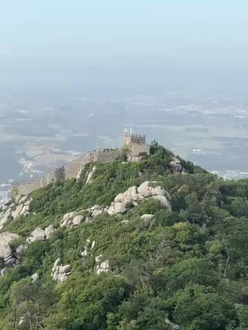 The video presents a series of three brief clips showcasing a historical site in a mountainous landscape, consistent with Sintra, Portugal.

The first clip (0:00-0:01) offers a wide, high-angle view of a medieval castle complex perched atop a green, rocky ridge. A stone wall with several towers, some adorned with flags, snakes along the hilltop. In the hazy distance, a valley with scattered buildings and urban development is visible under a bright, somewhat overcast sky.

The second clip (0:01-0:02) transitions to a closer view of a vibrant red building, likely part of a palace or a section of the castle. A large, dark, moss-covered rock dominates the immediate foreground, with a segment of a white-framed window visible on the building behind it.

The final clip (0:02-0:03) is an extremely close-up and out-of-focus shot of textured, mossy surfaces, probably the rock or ground near the red building, displaying a mix of green, brown, and grey tones.

No people or explicit text are visible in the video. The scenes are set during the daytime under hazy conditions, emphasizing the architectural grandeur and natural ruggedness of the location.