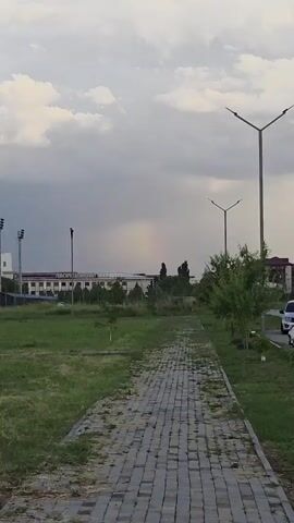 This media file captures an outdoor scene with a paved walkway in the foreground, bordered by green grass on the left and trees on the right. In the distance, a large, light-colored building with visible text is seen, flanked by trees and fencing. The sky is overcast with dramatic clouds, and a faint rainbow is visible above the building. The time of day appears to be late afternoon or early evening, given the lighting conditions.

The primary activity in the video is a person riding a bicycle along the paved walkway. The cyclist is dressed in dark clothing and is moving from right to left across the frame. The overall atmosphere is somewhat subdued due to the cloudy weather, but the presence of the rainbow suggests a recent or ongoing rain shower.

Notable details include the distinctive architecture of the distant building, which appears to be an institutional or commercial structure. The text on the building is partially obscured but seems to be in Cyrillic script. The presence of modern streetlights suggests an urban or suburban setting. The landscape is characterized by a mix of manicured grass and wilder vegetation, indicating a park or public space. The emotions evoked are calm and perhaps contemplative, with a hint of natural beauty from the rainbow.

The visible text on the building reads "ДВОРЕЦ КУЛЬТУРЫ", which translates to "Palace of Culture" in Russian. This identifies the building as a cultural center. The location context provided confirms this scene is in Argun, Russia.