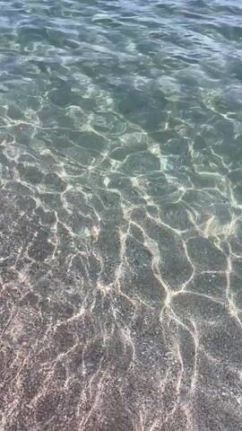 The video shows a close-up view of clear water with sunlight glinting off the surface. The water appears to be shallow, and the bottom is visible and appears to be sandy or rocky. The water is gently rippling, creating a mesmerizing pattern of light and shadow. There are no people or objects visible in the video. The environment appears to be a beach or a shallow body of water.