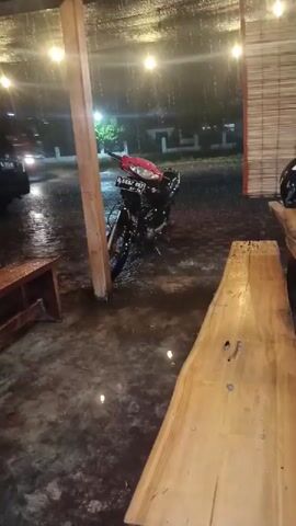 The video shows a motorcycle parked under a dimly lit shelter during a heavy rain.  The motorcycle is black with a red front and has a license plate that reads "B 6692 PAX". Behind the motorcycle, there is a "Pertamina" truck parked.  To the left of the motorcycle is a dark-colored car.  Inside the shelter, there is a small wooden bench to the left and a long, natural-wood table in the foreground on the right. The ground is wet and reflecting the string of lights hanging from the shelter's roof.  The overall scene suggests a roadside rest stop or outdoor café during a downpour.