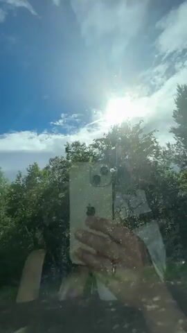 The video appears to be taken from inside a vehicle, looking out through the windshield or a side window. The sun is bright, creating lens flare and making it difficult to see some details clearly. The sky is blue with scattered white clouds. There are trees and lush green foliage visible outside the window. In the foreground, a person's hand is visible, holding a smartphone. The phone's screen is facing the camera, and a reflection shows the person taking the photo. The person is wearing a ring on their finger. The overall impression is a sunny day with a view of nature, possibly during a car ride.