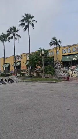 The video shows a wide shot of a long, one-story building with a yellow facade. The building appears to be abandoned or in disrepair, as it is covered in graffiti. Large palm trees are visible in front of and to the side of the building. In the foreground, there is a paved area that looks like a parking lot or road. There are several spherical sculptures in front of the building's entrance, which is covered by a dark-colored canopy. The sky is overcast.