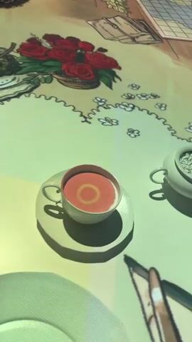 The video shows a close-up view of a table display with various objects arranged on it. The table is covered with a patterned tablecloth featuring illustrations of items like a cup of tea, a plate, a hand mirror, a fan, and food items. Text in German, possibly related to the items, is also visible on the tablecloth. The camera moves slightly, providing a shifting perspective on the objects and the text on the tablecloth. The light reflects off the surfaces of the objects, and the background appears blurred.