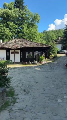 The video begins with a view of a cobblestone path leading towards a traditional Bulgarian house with a dark wooden facade and a stone tile roof. Lush green trees surround the house, and the sky is clear blue with some white clouds. As the camera pans, it reveals a small stream with stone walls on one side and more greenery. Further along, another house is visible, also with a stone tile roof and traditional architecture. The scene depicts a peaceful, rustic village environment with a focus on historical buildings and natural scenery. There are no people visible in the video.