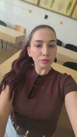 The video shows a woman with long, dark hair pulled back in a ponytail, wearing a brown t-shirt and a light-colored skirt. She is sitting in what appears to be a classroom or lecture hall, with desks visible in the background. The woman is taking a selfie video, and at different points she looks at the camera, winks, and makes a kissing face. Sunglasses are hanging from the collar of her shirt.