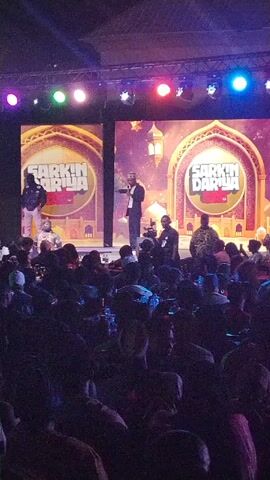 A crowded event is taking place indoors. A stage is set with two large screens displaying identical images of “Sarkin Dariya” against a background that suggests a Middle Eastern or Islamic theme. On stage, a man in a suit and cap is speaking into a microphone. Several other people are also on stage, including cameramen. A performer is walking towards the man in a suit. The audience is seated and many are holding up phones. The event seems to be well-lit and festive. The room is dim, with blue and yellow stage lights shining. The event is branded with “Maltina.”