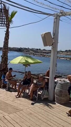 The video shows a sunny day at a beachside cafe. In the foreground, a wooden deck with white tables and chairs is visible. Several people are seated on chairs or standing. To the left, a tall palm tree stands next to a green umbrella. In the background, the sea is visible with many people swimming and sunbathing on the beach. The cafe appears to be open and operational, with a few patrons enjoying the atmosphere. The overall impression is one of a relaxed and pleasant day at the beach.
