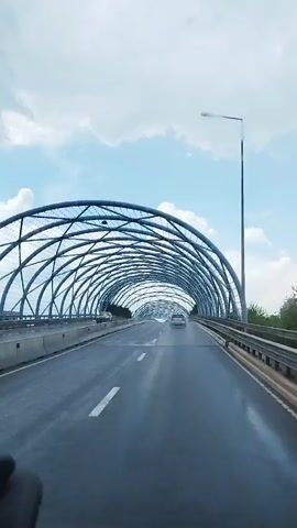 The video shows a view from inside a vehicle traveling on a highway. The highway is covered by an arched, lattice-like structure made of metal. The sky is visible through the open structure, with clouds present. There are several cars on the highway, and the road appears to be wet. The vehicle is moving forward and the scene is captured from the perspective of the driver or a passenger in the front seat.