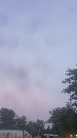 The video shows a cloudy sky, possibly dusk or dawn, with a few trees on the right side of the frame. A truck with "NQT" written on its side is visible in the distance, along with some lights and a white vehicle in the foreground on the left. The camera pans slightly to the right, revealing more of the trees and a dirt road. There are no people visible in the frame.