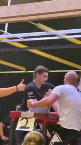 Here is a concise description of the video:

Two men are arm wrestling at a table in front of a crowd. A referee watches the match closely.  The table is labeled with the number two. The arm wrestling match occurs in an indoor sports hall.