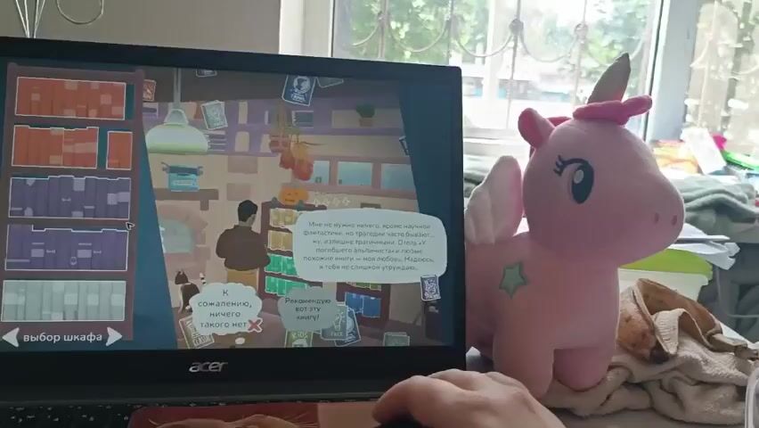 A person is playing a video game on a laptop. The game appears to be a visual novel or adventure game with a cartoonish art style. The screen shows a room filled with bookshelves and a character standing in the middle. Dialogue boxes are visible on the screen, indicating interaction or story progression. The laptop is placed on a surface, possibly a desk, with a computer mouse in front of it, operated by the person's hand. To the right of the laptop, a plush toy unicorn with pink wings and a pink horn is placed. The background is blurry but suggests an indoor setting with a window and natural light. The overall atmosphere is casual and relaxed.