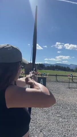 A woman with curly brown hair, wearing a black tank top and a grey baseball cap, is aiming a black bow and arrow at a target. Her arms are bent as she holds the bowstring taut. In the background, a green sign with a large white "A" is visible, along with several colorful targets shaped like balloons on stands. The scene takes place outdoors on a sunny day with a bright blue sky and scattered white clouds. The ground is covered with gravel. In the distance, there are trees and mountains. The woman releases the arrow, and it flies out of frame. The video then focuses on the targets.