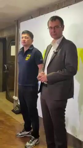 Two men stand in front of a white backdrop, likely indoors. The man on the left wears a dark blue polo shirt with a yellow emblem on the front, dark pants, and multicolored sneakers. The man on the right is wearing a light-colored button-down shirt under a dark brown suit jacket and trousers, glasses, and a microphone clipped to his shirt. The man on the right has his hands clasped in front of him. A dark wooden door and frame are visible on the left of the frame.