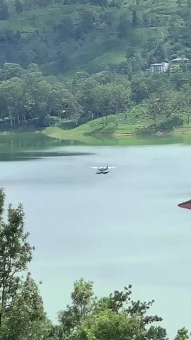 The media file captures a scenic view of a lake in Kuda Maskeliya, Sri Lanka. The main subjects are a seaplane and the natural landscape. The seaplane is a small, twin-engine aircraft with floats, positioned in the middle of the vast, calm lake. It appears to be in flight, possibly preparing for landing or taking off.

The setting is a lush, green environment. Rolling hills covered with dense trees form the backdrop, with some areas showing terraced plantations, likely tea estates, which are characteristic of Sri Lanka. The lake itself is a prominent feature, reflecting the surrounding greenery and the sky. The water appears tranquil, with gentle ripples.

In the distance, on the far side of the lake, there are hints of human structures, including what appears to be a building on a hilltop and some smaller structures near the shoreline. There are also a couple of yellow boats visible on the water, suggesting recreational activities or local transportation.

The lighting suggests it is daytime, likely late morning or afternoon, with diffused light that doesn't cast harsh shadows, implying overcast or slightly hazy weather. The overall atmosphere is serene and picturesque, highlighting the natural beauty of the location. There is no visible text in the media file.