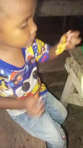 A young child, wearing a colorful t-shirt with cartoon characters and blue jeans, is sitting on a wooden surface and eating from a spoon. The child is bringing the spoon to their mouth, and then bringing food directly to their mouth with their hands. There are plates with food on a wooden table in the background. The environment appears to be outdoors at night.
