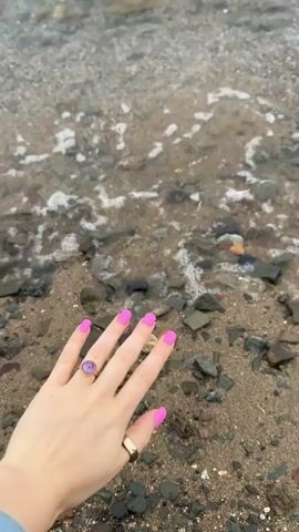 A close-up shot shows a person's left hand with bright pink fingernails and two rings, one with a purple stone, resting on a pebble and sand-covered shoreline. The edge of a body of water is visible, with small waves lapping against the shore. The hand is positioned near the water's edge, and the waves intermittently touch the tips of the fingers. The lighting suggests an overcast day or early morning/late evening.