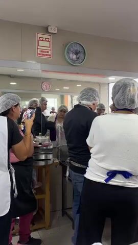 A group of people, most wearing hairnets, are gathered in a kitchen or food preparation area. Several individuals appear to be participating in a cooking class or demonstration. In the foreground, a person in a white shirt and black pants is visible from behind, wearing a blue apron. To their left, a person in a pink apron is holding a smartphone, seemingly taking a picture or recording. In the middle ground, a man in a black shirt and blue apron is interacting with others, some of whom are also wearing aprons and hairnets. A woman in a black jacket and hairnet is holding a red object, possibly a food item or utensil, and is looking at someone. Another woman in a pink apron is standing beside her. The environment includes stainless steel equipment, bowls, bananas, eggs, and various food containers, suggesting active food preparation. A clock is visible on the wall, as well as a red and white emergency light sign. The overall scene depicts a collaborative or educational activity centered around food.