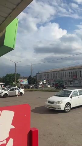 This is a vertical video clip shot from a low angle, possibly from the perspective of someone standing at a bus stop or near a shop. The scene is an outdoor street in Yegorlykskaya, Russia, under a cloudy sky that suggests it might be late afternoon or early evening, with a hint of a storm approaching.

In the foreground, a bright red object, likely a sign or part of a building structure, partially obscures the view on the left. Moving into the midground, several cars are visible on a paved road. A white car is prominently featured on the right side of the frame, with its license plate reading "Y207OE 123 RUS". Another white car is parked on the left, and a person in a light-colored top and shorts is standing next to it, looking towards the street. There are other vehicles, including a dark SUV and a few sedans, parked or driving in the background.

The background reveals a town setting. A tall cell tower stands prominently against the dramatic, cloud-filled sky. To the right of the tower is a light-colored building with multiple windows and a sign that reads "ПРОДУКТЫ" (PRODUCTS) and "МЯСНАЯ ФЕРМА" (MEAT FARM). Other signs in Russian are visible on buildings and poles, including what appears to be a bus stop sign with the word "ТАКСИ" (TAXI) and numbers indicating a distance of "12.5 м". A large billboard on the left features an image of a woman and text including "ПОБЕДА НАШИ" (VICTORY IS OURS) and numbers that appear to be prices or contact information.

The overall atmosphere is that of a typical day in a Russian town, with everyday activities like driving and walking taking place. The cloudy sky adds a touch of drama to the scene.