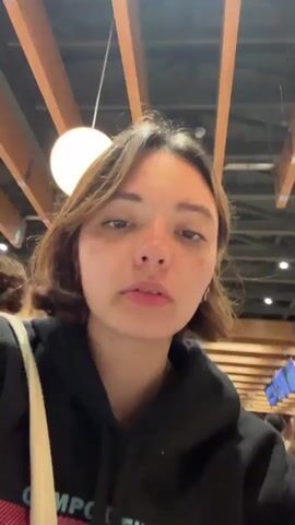 The video shows two young adults, a female on the left and a male on the right, taking a selfie in what appears to be a crowded indoor public space, possibly a food court or marketplace, with a blurred background of other people and structural elements of the building. The female has short brown hair and is wearing a black hoodie with pink accents. She is looking at the camera with a neutral expression. The male has dark hair and a beard, and is wearing a light-colored t-shirt. He is leaning in towards the female and making various facial expressions, including sticking out his tongue. The lighting suggests it is daytime.