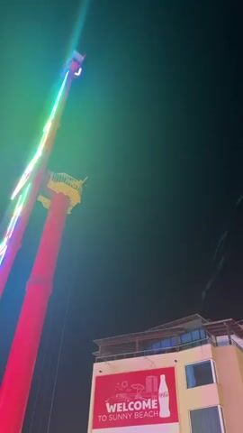 The video shows a tall amusement park ride at night. The ride, which appears to be a pendulum-style swing, has bright lights that change color from green to blue to purple. At the top of the ride, a small gondola is visible. Below the ride, a building with a sign that says "WELCOME TO SUNNY BEACH" can be seen. The sky is dark, indicating it is nighttime. The camera is positioned looking up at the ride, creating a dramatic perspective.