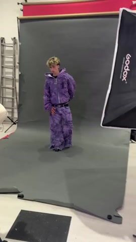 A young man is standing in front of a gray backdrop in a photography studio. He's wearing a purple outfit that appears to be distressed or ripped. The outfit consists of a hooded jacket and pants. The backdrop is part of a photography setup, which includes a red roll at the top, and a large softbox light stand is visible to the right. There's a ladder in the background to the left, along with other photography equipment. A person is partially visible in the foreground, suggesting someone is taking photos or videos of the young man. The room has a light-colored floor and good lighting. Club music is playing in the background.