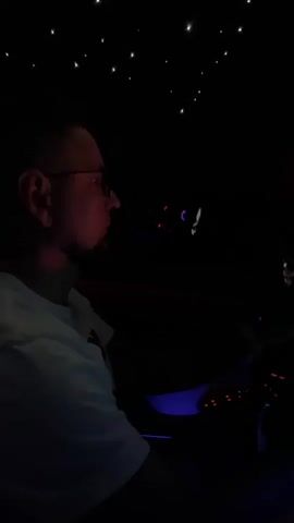 A person wearing glasses and a white t-shirt is seen from the side, sitting in the driver's seat of a car at night. The car's interior is dimly lit, with ambient blue and red lights illuminating the dashboard and steering wheel. The ceiling of the car appears to have a starry effect, with small lights resembling stars. The person's hands are visible, one on the steering wheel and the other near the center console, suggesting they are driving or interacting with the car's controls. The dashboard displays illuminated information, and a navigation screen is visible to the right. The overall atmosphere is dark and futuristic, likely due to the car's interior lighting.