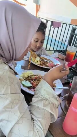 The video shows two people, an adult and a young child, sitting at a table and eating. The adult is wearing a light-colored patterned top and a hijab. The child is wearing a light-colored top and has a yellow hair accessory. They are both eating plates of food that appear to contain noodles, vegetables, and a sauce. There are drinks on the table, including iced tea and a bottle of sauce. The table is outdoors on a patio or balcony with a black metal railing. The surrounding environment includes buildings with white and orange exteriors. The camera focuses on the food and the actions of eating.
