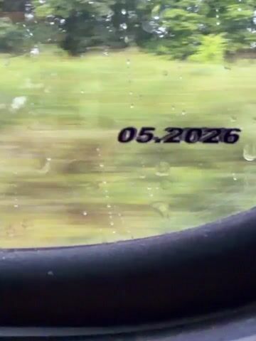 A close-up shot shows a date stamp, 05.2026, on a window, possibly of a vehicle. The background is blurred, suggesting motion, and the scenery appears to be green and outdoors, with some raindrops visible on the window. The framing of the shot focuses on the date stamp and the edge of the window's frame.